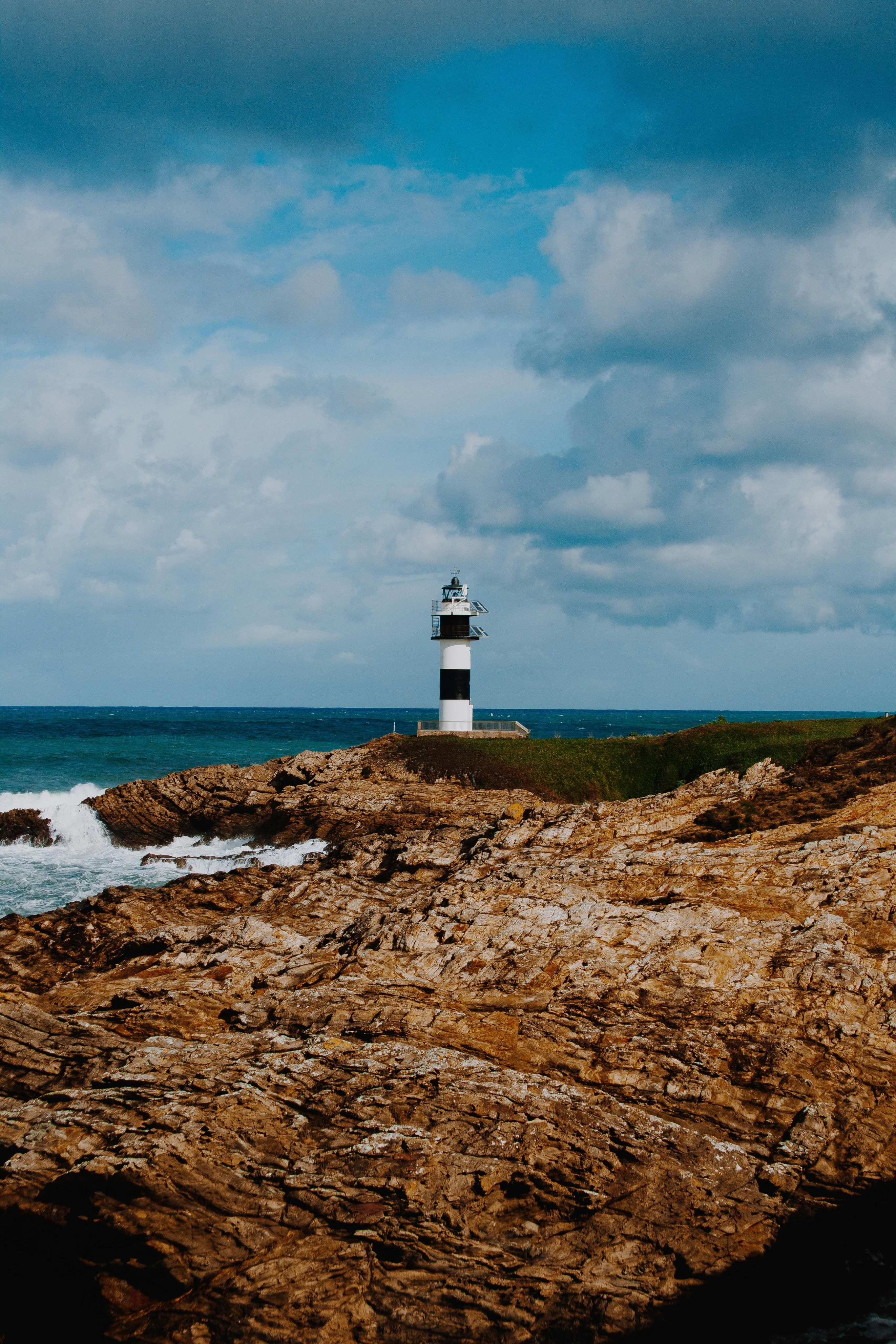 White lighthouse with a black stripe in the middle, standing on a rocky cliff by the sea, with the ocean horizon and cloudy sky in the background.
